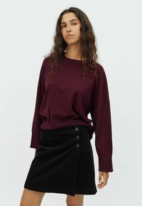 Pull-over en maroon à la coupe ample associé à une jupe en velours côtelé noire avec trois boutons sur le côté et un ourlet diagonal.