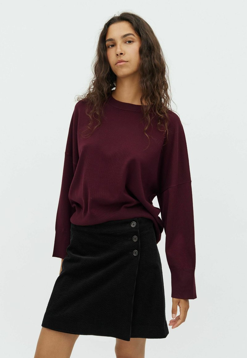 Pull-over en maroon à la coupe ample associé à une jupe en velours côtelé noire avec trois boutons sur le côté et un ourlet diagonal.