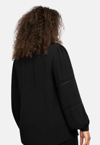 Černá blůza s dlouhými nabíranými rukávy, která má průhledné výřezy na rukávech, hladký povrch a uvolněný střih.