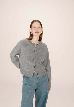 Cardigan gris avec un col rond, fermeture à boutons et ceinture à côtes. Porté par-dessus un jean large bleu, avec une texture tricot douce.