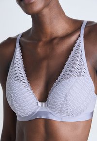 Bralette en dentelle grise avec un design en V profond. Présente des bords festonnés, un sous-bande lisse et un fermoir central en métal.