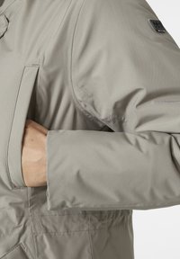 Chaqueta de manga larga color gris claro con tejido texturizado, que presenta un bolsillo frontal y un acento de marca en el hombro.