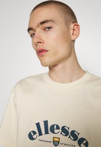 Camiseta de color beige claro con texto de marca en azul oscuro. Diseño de cuello redondo con una textura suave. Mangas cortas y un ajuste relajado.