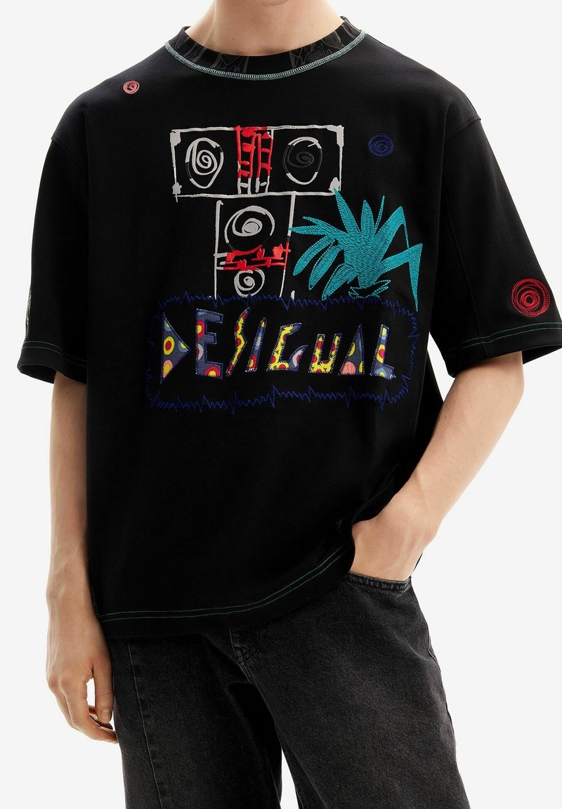 Camiseta negra de gran tamaño con formas abstractas coloridas, una planta bordada en azul y la palabra "DESIGUAL" en letras multicolores.