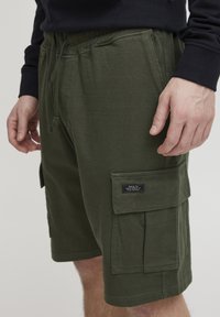 Grønne bomullscargo-shortser med elastisk midjebånd, sidelommer og en klaffelomme med et merke. Myk og pustende tekstur.