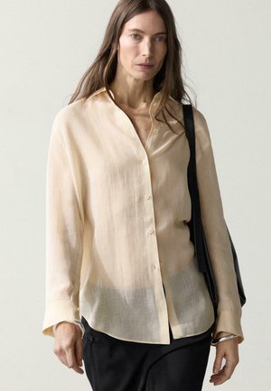 Femme portant une blouse ample beige clair boutonnée et un pantalon noir, avec un sac à bandoulière noir, debout devant un fond uni.