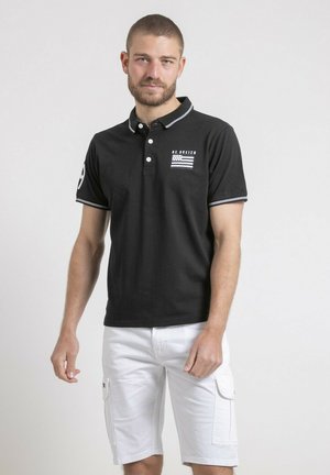 Homme aux cheveux courts et barbe, portant un polo noir avec logo et un short cargo blanc, debout devant un fond clair uni.