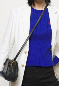 Blå kabelstickad tröja under en vit kavaj, kombinerad med svarta byxor. En liten svart handväska med guldig dragkedja och rem bärs tvärs över kroppen.