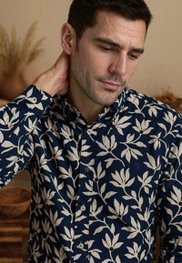 Marineblauwe shirt met wit bloemenpatroon, met knopsluiting en subtiele logo-detail. Katoenen stof met een gladde textuur en een getailleerde stijl.