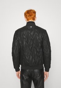 Veste bomber noire matelassée avec un col montant, des poignets et un ourlet côtelés. Présente un motif texturé et un petit logo à l'arrière.