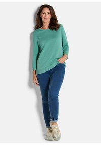 Frau, die einen strukturierten, türkisfarbenen Pullover, dunkelblaue, hochgekrempelte Jeans und beigefarbene Sneakers trägt, vor einem weißen Hintergrund steht.