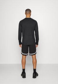 Muž stojící zády, oblečený v černém tričku s dlouhým rukávem, černých kraťasech s bílým lemem, černých ponožkách a černých sportovních botách.