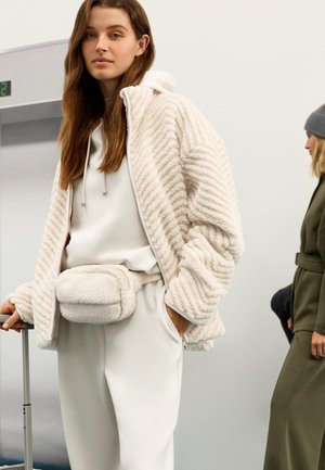 Jakna z zadrgo v kremni barvi z teksturo in usklajeno torbico za pas. Jakna ima cikcak vzorec in je kombinirana z lahko kapuco in hlačami.