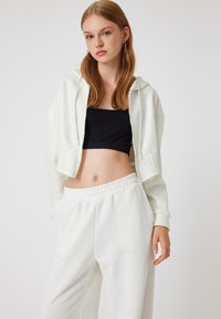 Felpa bianca con zip, tagliata, con cappuccio, top corto nero a costine sotto e pantaloni della tuta bianchi. Tessuto morbido e vita elastica.
