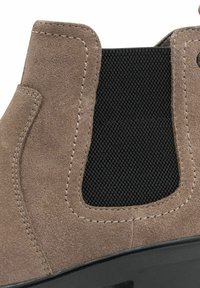 Cizmă scurtă din suede maro, cu panou elastic negru pe lateral, detalii cusute și talpă din cauciuc, având o textură netedă și un design modern.