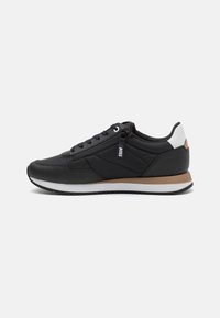 Zapatilla deportiva negra con parte superior de cuero y textil liso. Cuenta con una suela intermedia blanca y detalles en color beige, incluyendo un logo en el lateral.