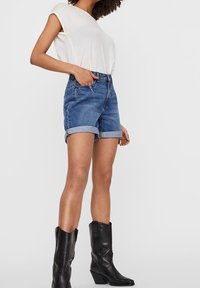 Mujer con una blusa sin mangas blanca, pantalones cortos de mezclilla azul arremangados y botas de vaquero negras hasta medio pantorrilla, de pie con una mano en el bolsillo.