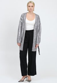 Silberner Pailletten-Cardigan mit gerippter Textur, offenem Vorderteil und langen Ärmeln, kombiniert mit hochtaillierten schwarzen Hosen und gemusterten Absätzen.