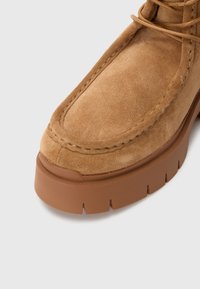 Botín de ante marrón hasta el tobillo con una suela de goma gruesa y texturizada. Presenta un diseño cosido a lo largo del borde y detalles de cordones en la parte superior.