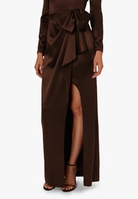 Długa, brązowa satynowa sukienka z dużą kokardą w talii i rozcięciem do uda, zestawiona z czarnymi sandałami na obcasie, eksponując jedną nogę.