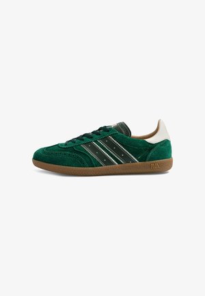 Baskets en daim vert avec des rayures noires et blanches, lacets noirs, languette blanche au talon, semelle marron avec une accroche texturée et petit logo.