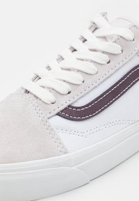 Baskets blanches et beiges avec lacets blancs, bande latérale marron texturée, et semelle en caoutchouc blanche sur fond blanc.