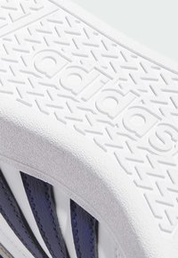 Weiße Gummisohle mit strukturiertem Profil und geprägt "adidas"-Logo. Verfügt über dunkelblaue Ledestreifen für Halt und Design.