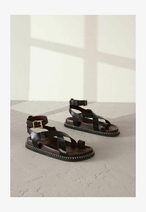 Juodos odinės šlepetės su kryžminiais dirželiais ir atviromis pirštų dalimis. Yra kulkšnies dirželis su sagtimi ir kontrastingai siuvinėtas padas.