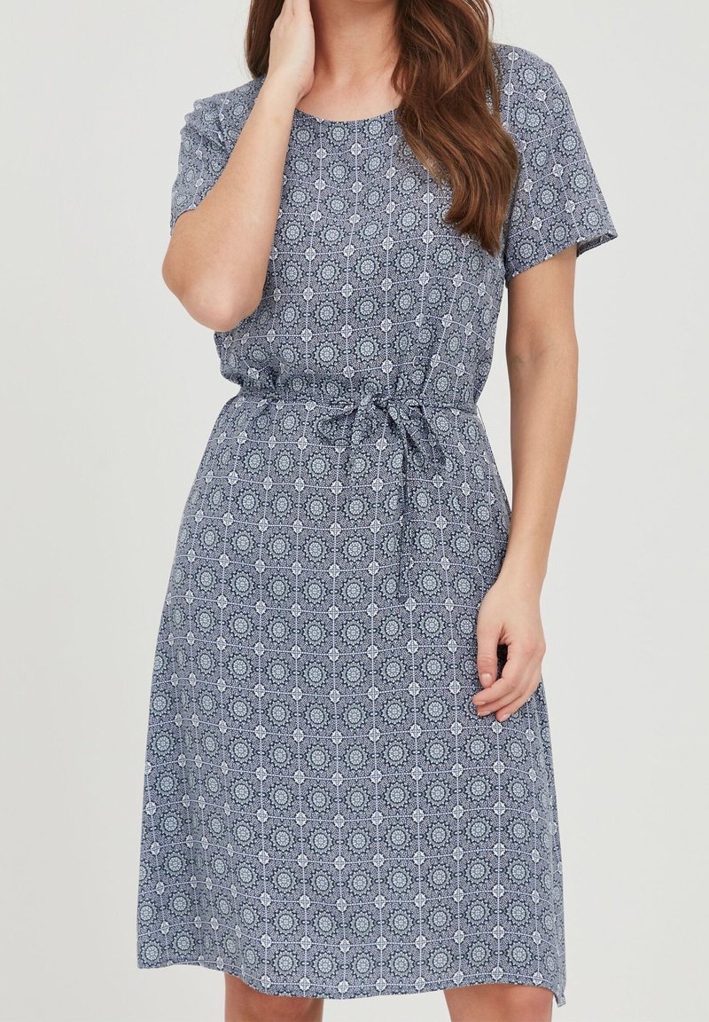 Femme portant une robe à manches courtes, longueur genou, avec un motif géométrique bleu et blanc et une ceinture nouée à la taille.