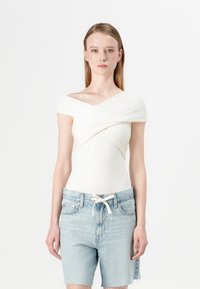 Bijela off-shoulder majica s dizajnom uvijenog prednjeg dijela, izrađena od mekog materijala, u kombinaciji sa svijetlo plavim trapericama s elastičnim pojasom.