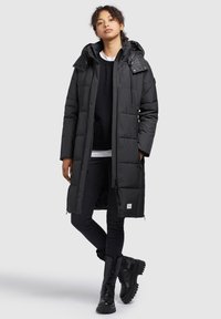 Black padded coat featuring a hood, zip closure, and side pockets. Worn over a black sweater and paired with black skinny jeans and boots.