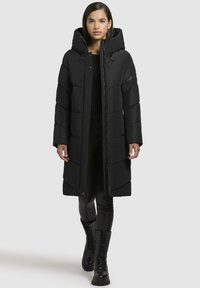 Ilgas juodas pūkų paltas su kapišonu, turintis užtrauktuko ir mygtuko uždarymą, dideles kišenes ir siuvinėtą dizainą. Dėvimas su juodomis legginsomis ir batais.