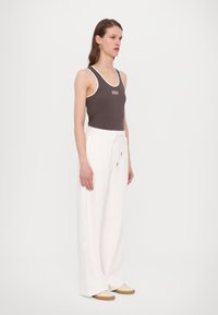 Brun ribbet tanktop med hvit kant og "MM"-logo; sammen med løse hvite bukser med snøring og side-seam detaljer.