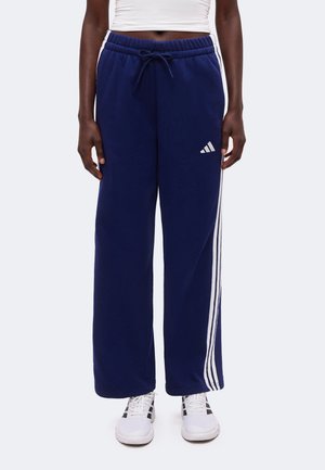 Personne portant un pantalon de survêtement Adidas bleu marine avec des bandes blanches et un logo, des chaussures de sport blanches et un top court blanc.