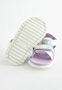 Lila sandal med ett glänsande, iridescerande rem, justerbar stängning och en strukturerad vit gummisula med parallella spår.