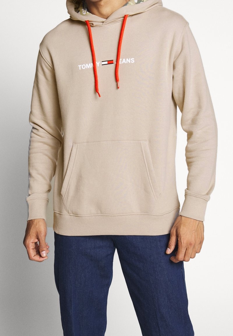 Sudadera con capucha beige con cordones naranja contrastantes y un bolsillo canguro frontal. Presenta el logo "TOMMY JEANS" en blanco en el pecho.