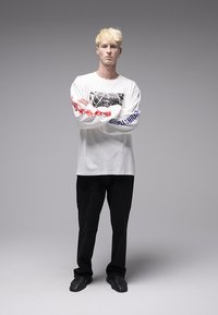 Λευκό πουκάμισο με μακριά μανίκια, μαύρο γραφικό σχέδιο και κόκκινο κείμενο σε ένα μανίκι, συνδυασμένο με μαύρο παντελόνι και μαύρα παπούτσια.