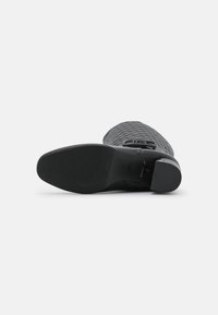 Bota de caña alta negra texturizada tumbada de lado, mostrando la suela suave y el tacón bajo con un sutil patrón de agarre.