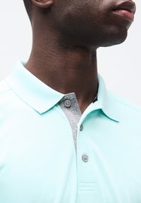 Κοντινό πλάνο στο λαιμό και το κάτω μέρος του προσώπου ενός άνδρα που φοράει ανοιχτό μπλε πόλο με γκρι κουμπότρυπες και γιακά.