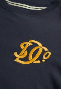 Ciemnogranatowa tkanina z wyhaftowanymi złoto-żółtymi stylizowanymi literami tworzącymi splecione logo w pobliżu zaokrąglonego dekoltu z białym paskowanym wykończeniem.