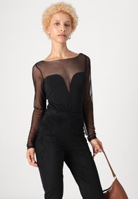 Crna bodysuit s prozirnim dugim rukavima, dekolteom u obliku srca i crnim hlačama visokog struka. Model drži smeđu torbu.