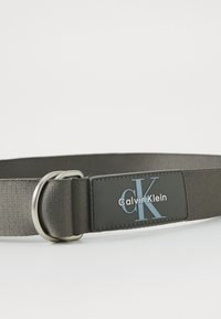 Calvin Klein belt in gray fabric with a smooth texture, featuring a metal ring buckle and a black logo patch with blue accents.