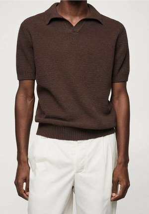 Mężczyzna w brązowej, krótkorękawowej dzianinowej koszulce polo i białych spodniach, stojący na tle prostego jasnego tła.