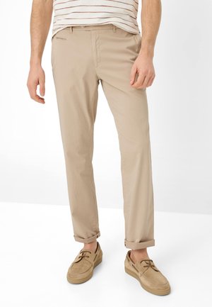 Man die beige opgerolde chino's draagt, bruine veterschoenen en een wit overhemd met bruine horizontale strepen, staande tegen een witte achtergrond.