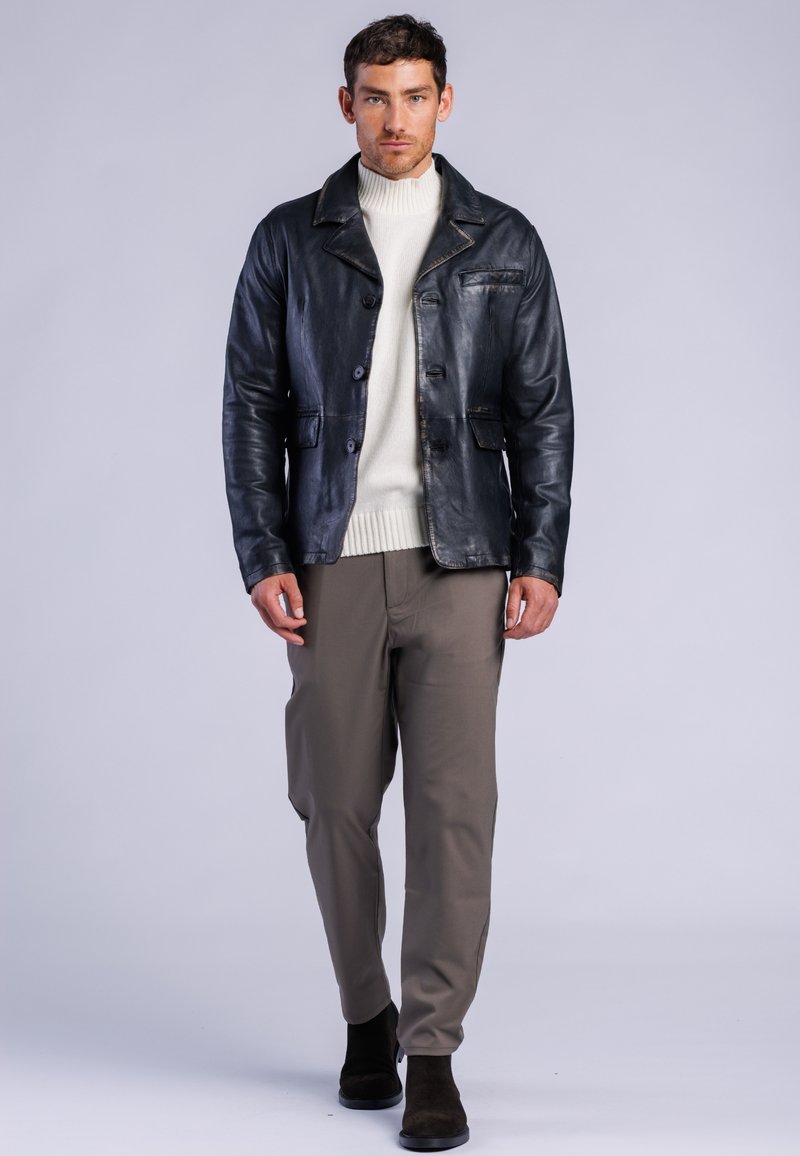 Hombre con una chaqueta de cuero negra, suéter de cuello alto blanco, pantalones grises y botas negras caminando hacia adelante sobre un fondo claro y liso.