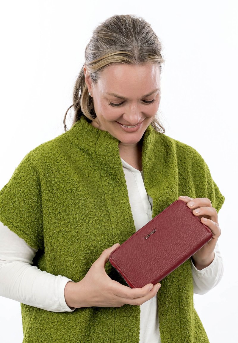 Mujer con chal texturizado verde sosteniendo y mirando hacia abajo una cartera de cuero roja con cremallera y nombre de marca en relieve.