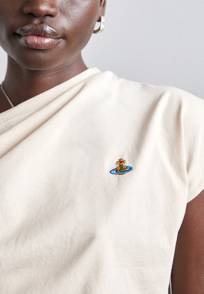 Tricou din bumbac crem cu un decolteu rotund. Prezintă un design brodat colorat al unei broaște țestoase pe un fundal circular albastru, situat pe piept.