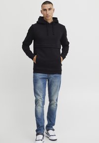 Sudadera negra con bolsillo canguro, capucha con cordón y una banda texturizada horizontal. Combinada con jeans de color azul claro y zapatillas blancas.