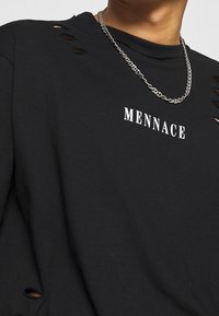 Černé bavlněné tričko s žebrovaným kulatým výstřihem, bočními rozparky a výřezovými detaily. Přední potisk v bílé barvě nese nápis "MENNACE". Stříbrný řetízek.