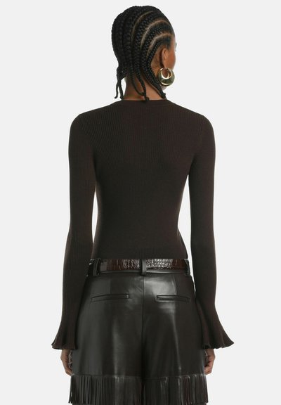 Top lungo a maniche ribbed marrone con polsini svasati, abbinato a pantaloni corti in pelle nera con dettagli in frange e una cintura marrone testurizzata.